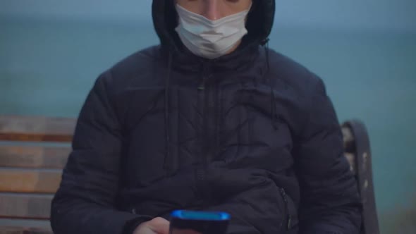 A Man in a Medical Mask Sits on a Bench Near the Sea and Looks at the Phone