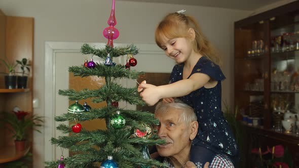 Girl Kid with Senior Grandpa Decorating Artificial Christmas Tree with Ornaments and Toys at Home