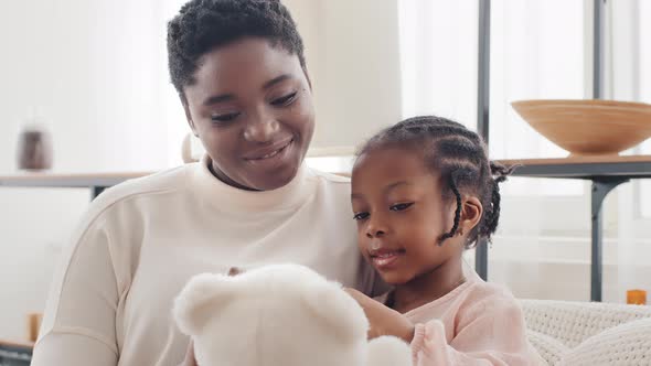 Afro American Mother with Little Daughter Girl Sitting on Sofa at Home Talking Holding Teddy Bear