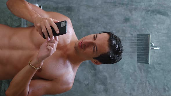 Vertical Video Portrait of Smiling Young Man Stands in the Bathroom Near the Shower and Using App