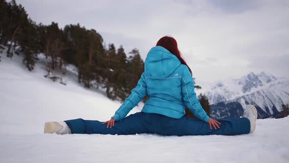 Girl in Skiing Equipment Sits in Splits Against Backdrop Mountain Winter