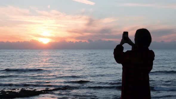 Dark Silhouette of young girl taking photos of sunset at wavy sea