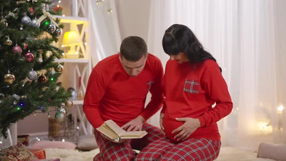 Happy Smiling Man Reading Book and Kissing Pregnant Belly of Smiling Woman