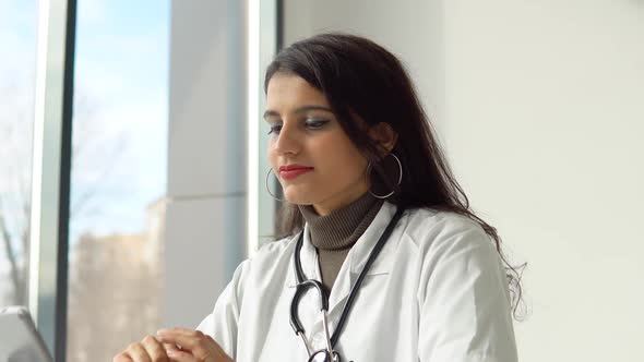 Indian Female Doctor in a White Coat with a Stethoscope Conducts an Online Consultation in Her