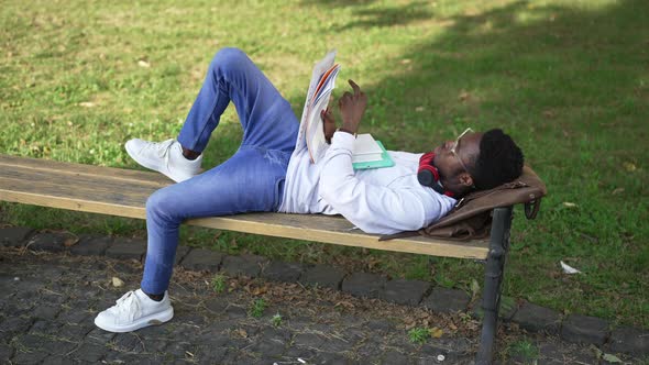 Wide Shot Relaxed Confident African American Student Lying on Bench Outdoors Learning Material