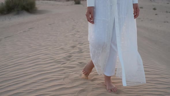 A Woman in a White Dress Barefoot is Walking Along the Desert Sand