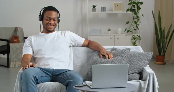 African Man Male Student Listens to Music with Headphones Sibging Sitting at Home By Computer