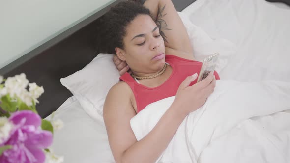 Cute African American Woman Just Woke Up, She Lying in the Bed Texting on Cellphone. Happy Girl