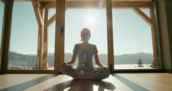Meditating Woman in Pajamas Sits in Lotus Pose By Big Window in Morning at Home