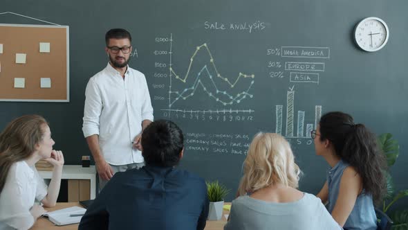 Cheerful Young Businessman Discussing Project with Colleagues and Pointing at Chalkboard in Office