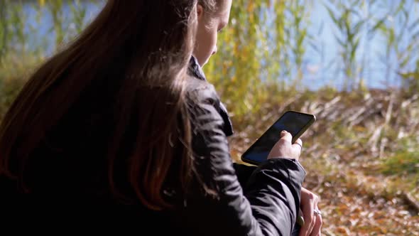 Young Woman Reading Text Message in a Smartphone Sitting on Fallen Dry Leaves