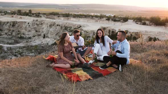 Four Friends Cheering with Beer and Smiling While Sitting on the Blanket at Picnic