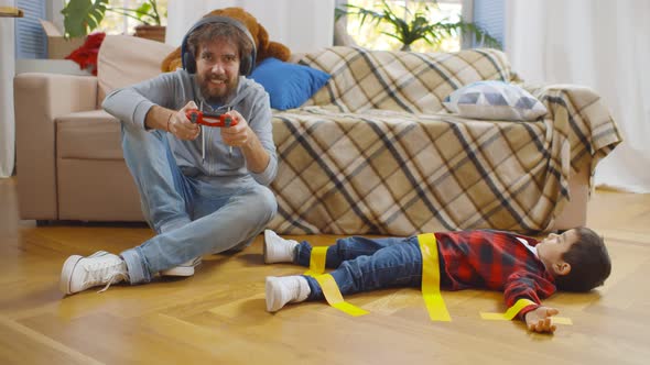 Father in Headphones Playing Video Game with Console and Disobedient Son Tied with Tape on Floor