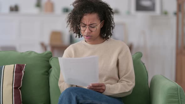 Young African Woman with Documents Reacting To Loss on Sofa 