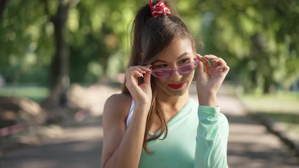 Charming Slim Young Caucasian Woman with Ponytail in Pink Sunglasses Posing Outdoors in Sunny Park