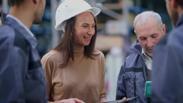 Portrait of Beautiful Cheerful Woman in Hard Hat Talking with Men Smiling Standing in Industrial