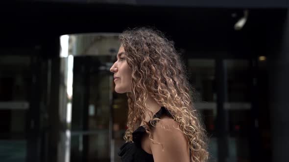 a Young Woman Walks Down a City Street and Adjusts Her Curly Hair. Side View