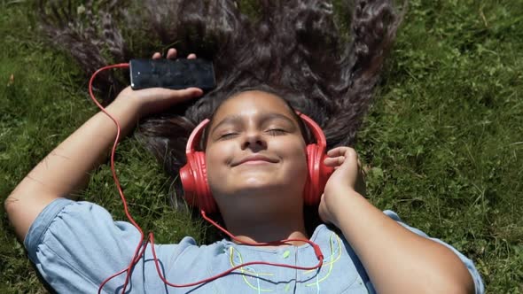 Attractive Girl with Long Hair Lies on the Grass in the Park and Listens to Music in Red Headphones