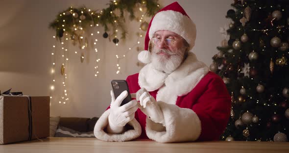 Santa Claus Sitting at a Table Near a Christmas Gift Box in Craft Wrapping Paper and Using Mobile