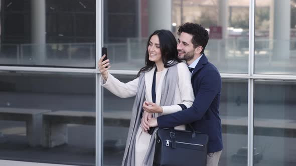 Business couple taking selfie, embracing in front of modern building