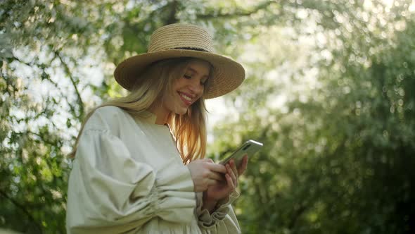 Portrait of Blonde Woman in Hat Enjoy Takes Pictures at on Smartphone Device of White Apple Blossoms