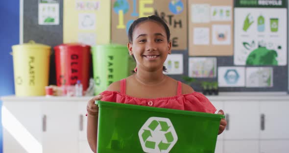 Mixed race schoolgirl smiling, holding recycling bin, standing in classroom