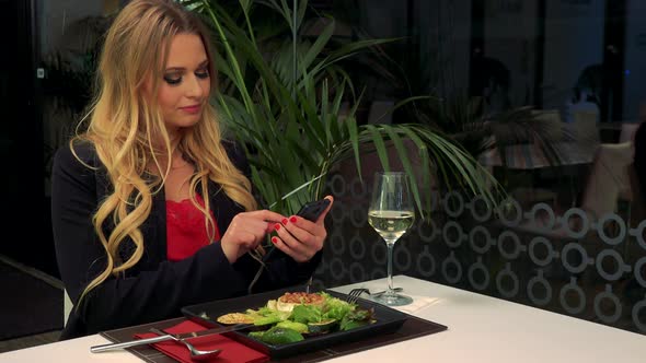 A Young, Attractive Woman Sits at a Table in a Restaurant and Works on a Smartphone