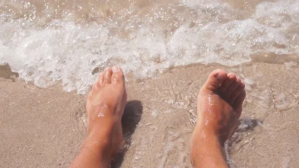 Closeup of Men's Legs Lying on the Sea Coast Small Waves Hitting Their Feet on the Beach in Slow