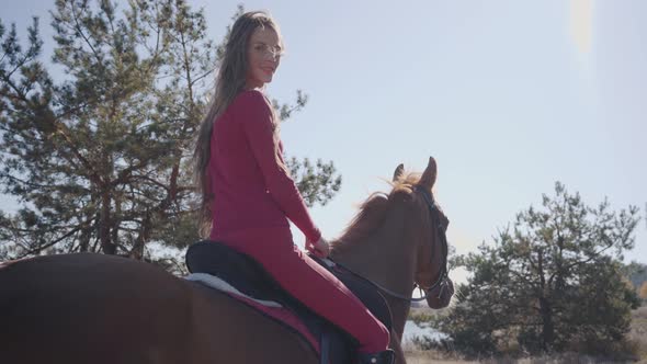 Back View of a Smiling Caucasian Female Equestrian Sitting in Sunlight on the Horseback and Riding