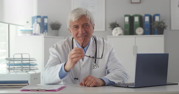 Portrait of Aged Doctor Sitting at Desk and Smiling at Camera in Clinic Office