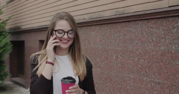 Joyful Girl Walks with Cup of Beverage in Hand and Talking on Phone with Smile