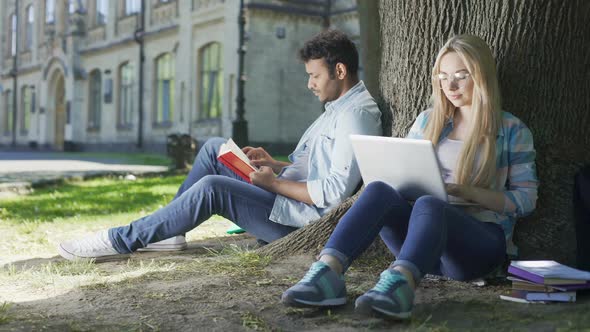 Male With Book, Female With Laptop Under Tree Looking at Each Other and Smiling