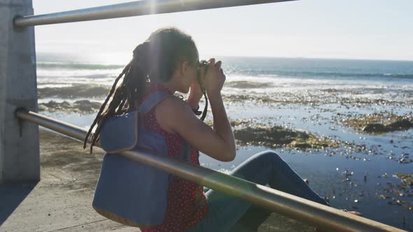 African american woman sitting and photographing on promenade by the sea