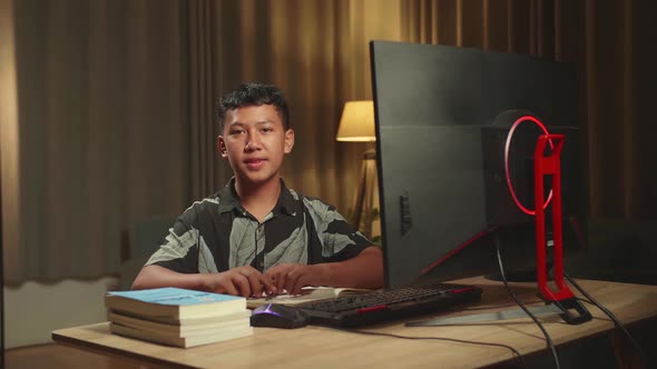 Asian Boy Learning Online With Desktop Computer From Home, He Turns And Smiles Into The Camera