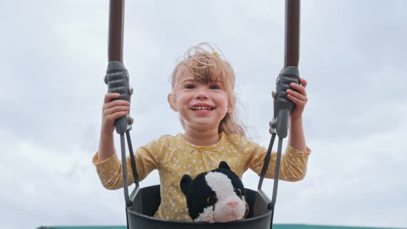 Happy Little Girl Lwith Toy Cat Smiling at Camera While Swinging at Toddler Swing