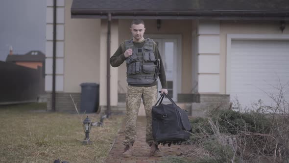 The Young Soldier in Military Closes with Big Bag Standing in Front of the House