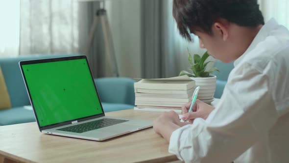 Asian Man Student Look At Green Screen Computer And Write In The Notebook While Studying Online