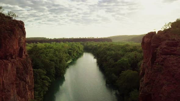 Drone flying straight out of a gorge between two large red rock walls following a river surrounded b