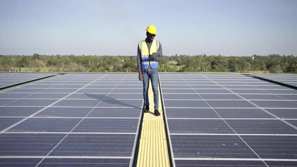 4K African man engineer using digital tablet maintaining solar cell panels on building rooftop.