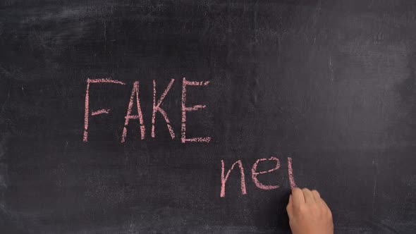 A man's hand writes the phrase FAKE NEWS in red chalk on a black chalk board