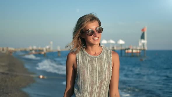 Smiling Young Girl in Trendy Sunglasses Walking on Beach at Sunset Enjoying Freedom