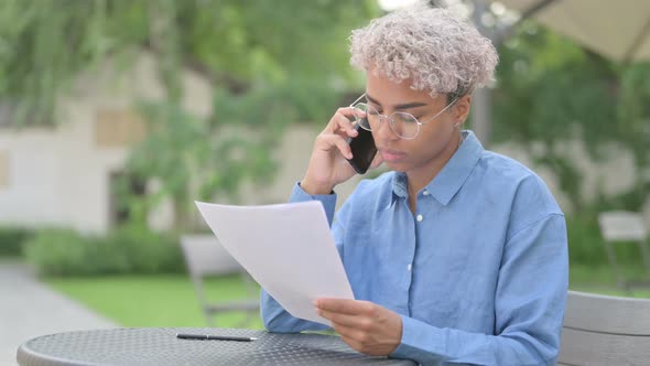 Young African Woman Talking on Phone Reading Documents in Outdoor Cafe