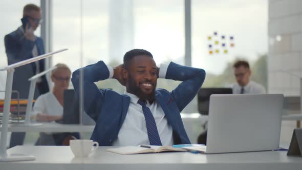 Happy Afro Businessman with Hands Behind Head Smiling Resting at Desk in Office.