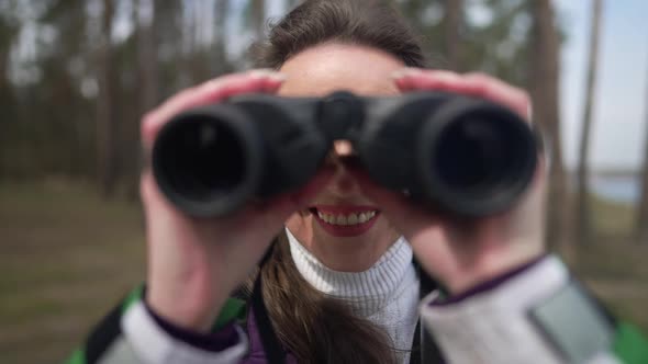 Front View Smiling Woman Using Binoculars Looking at Camera Standing in Forest