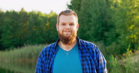 Portrait of a Smiling Young Man with a Red Beard Outdoors in a Forest Near a Lake