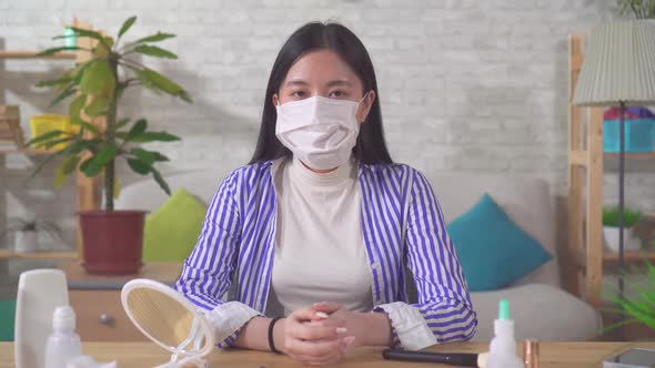 Young Asian Woman Puts Medical Mask While Sitting Home Table Looks Camera