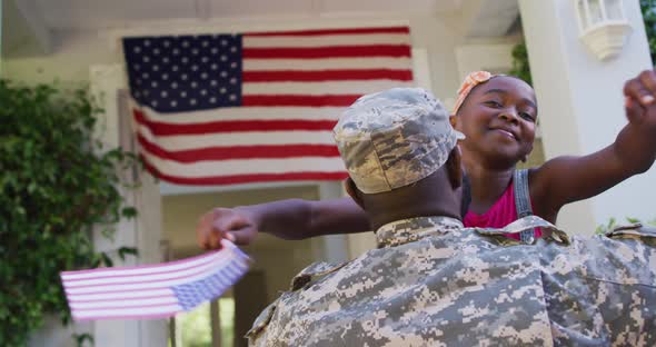 African american soldier father hugging smiling daughter in front of house with american flag