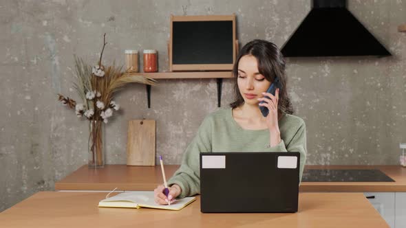 Girl Sitting at Laptop in Her Living Room Talking on Phone and Writing Notes