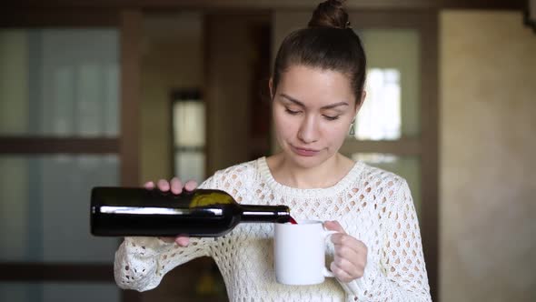 Girl pours red wine into a mug and drinks, looking out the window in the kitchen