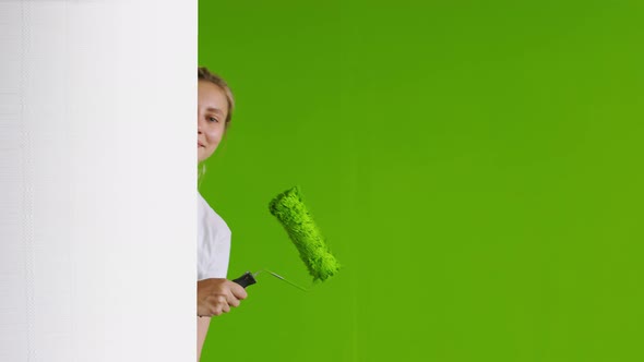 A Young Happy Woman Looks Out From Behind a White Wall with a Roller in Green Paint Backdrop a Green
